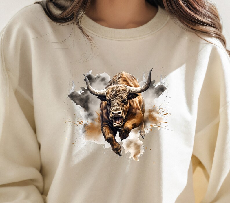 May include: A white sweatshirt with a brown and black bull illustration. The bull is charging forward with its mouth open and horns pointed forward. The bull is surrounded by a watercolor-style background of brown and gray splatters.