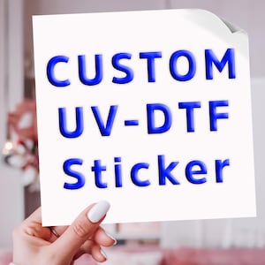 May include: A square white sticker featuring the text CUSTOM UV-DTF Sticker in bold, three-dimensional blue lettering. The sticker has a slight curl at the top right corner, showcasing a clean, professional design for custom labeling applications.