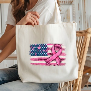 May include: A white tote bag with a pink ribbon and an American flag design. The flag is painted in a distressed style with pink hearts and stars. The ribbon is tied in a bow and has a leopard print pattern.