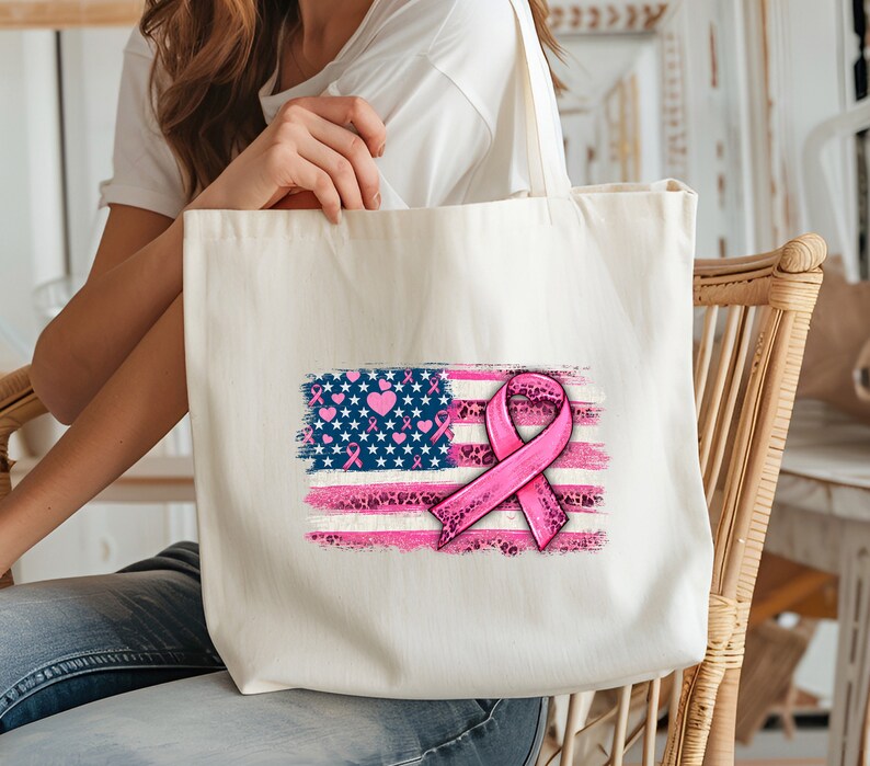 May include: A white tote bag with a pink ribbon and an American flag design. The flag is painted in a distressed style with pink hearts and stars. The ribbon is tied in a bow and has a leopard print pattern.