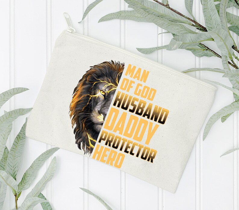May include: A white canvas zippered pouch with a lion graphic and the text "Man of God Husband Daddy Protector Hero" in yellow.