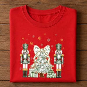 May include: A bright red crew neck t-shirt featuring a graphic print of two green nutcracker figures standing on either side of a decorated Christmas tree with a large bow on top and gift boxes at the base, surrounded by gold and white snowflakes.