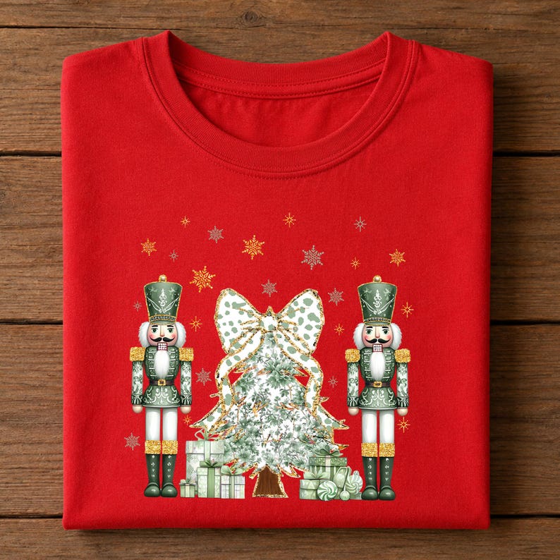 May include: A bright red crew neck t-shirt featuring a graphic print of two green nutcracker figures standing on either side of a decorated Christmas tree with a large bow on top and gift boxes at the base, surrounded by gold and white snowflakes.