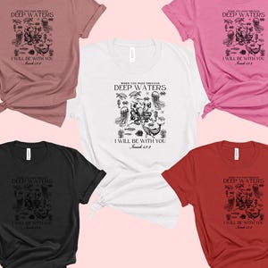 May include: A collection of short-sleeved t-shirts in various colors, including white, black, pink, red, and mauve. Each shirt features a black graphic with the text "WHEN YOU PASS THROUGH DEEP WATERS, I WILL BE WITH YOU, Isaiah 43:2."