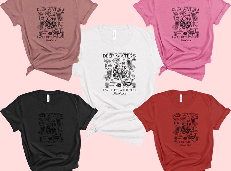 May include: A collection of short-sleeved t-shirts in various colors, including white, black, pink, red, and mauve. Each shirt features a black graphic with the text "WHEN YOU PASS THROUGH DEEP WATERS, I WILL BE WITH YOU, Isaiah 43:2."
