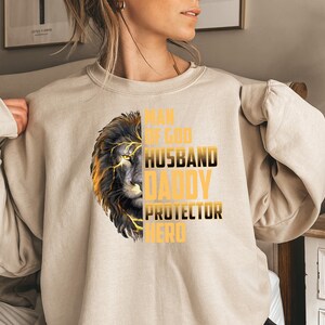 May include: A beige sweatshirt with a lion graphic and the text "Man of God, Husband, Daddy, Protector, Hero" in gold lettering.