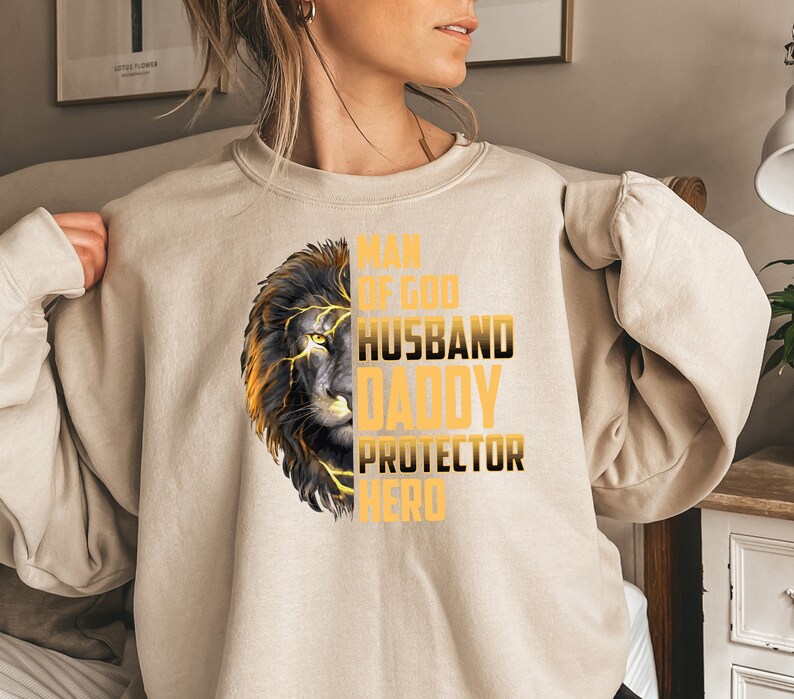 May include: A beige sweatshirt with a lion graphic and the text "Man of God, Husband, Daddy, Protector, Hero" in gold lettering.