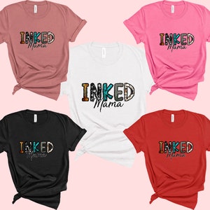 以下が含まれることがあります： ピンクの背景に、5色のTシャツが並んでいます。それぞれのTシャツには、カラフルな文字で「INKED Mama」と書いてあります。Tシャツはすべて折り畳んで並べてあります。