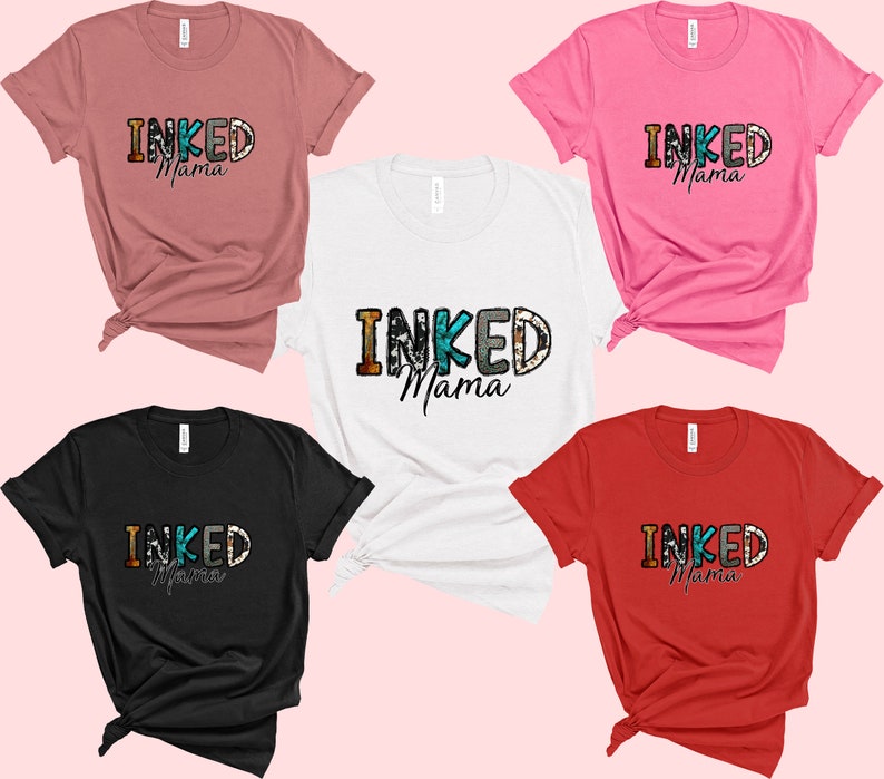 以下が含まれることがあります： ピンクの背景に、5色のTシャツが並んでいます。それぞれのTシャツには、カラフルな文字で「INKED Mama」と書いてあります。Tシャツはすべて折り畳んで並べてあります。