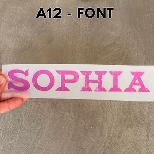 May include: A rectangular transfer sheet featuring the name SOPHIA in a bold, pink, distressed-texture font. The text is printed on a clear, semi-transparent backing material. The label A12 - FONT is visible at the top of the image.