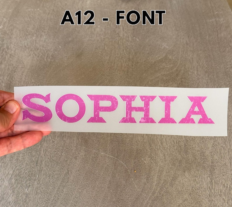 May include: A rectangular transfer sheet featuring the name SOPHIA in a bold, pink, distressed-texture font. The text is printed on a clear, semi-transparent backing material. The label A12 - FONT is visible at the top of the image.