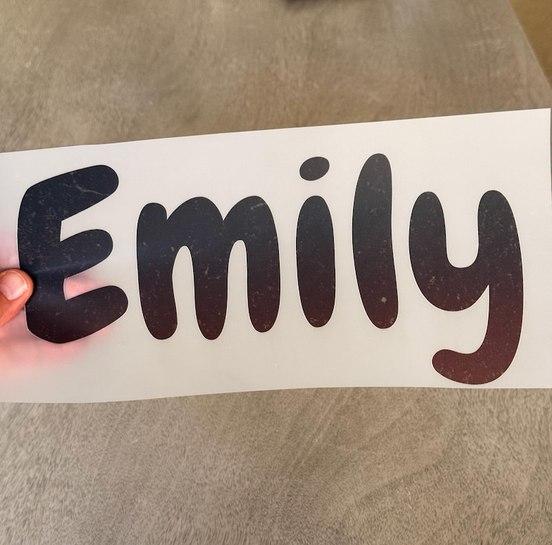 May include: A custom vinyl decal featuring the name Emily in a rounded, bold font. The lettering displays a dark gradient effect that transitions from black at the top to a deep reddish-brown at the bottom, set against a white backing sheet.