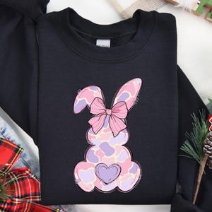 May include: Black sweatshirt featuring a pink and purple bunny with a pink bow. The bunny has a heart on its chest.