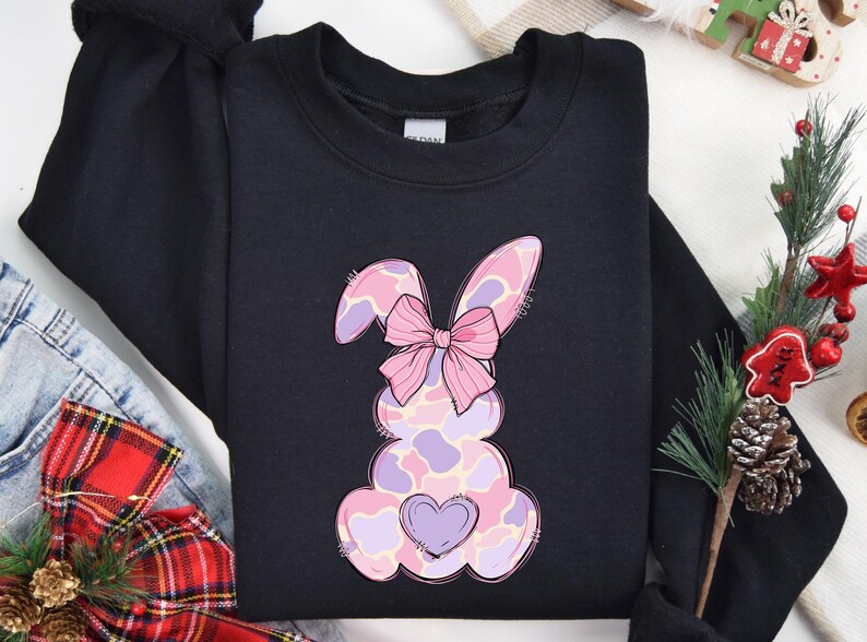 May include: Black sweatshirt featuring a pink and purple bunny with a pink bow. The bunny has a heart on its chest.