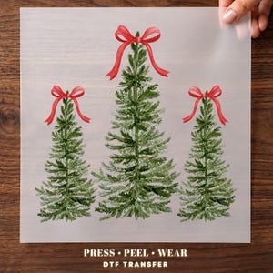 May include: A DTF transfer sheet featuring three green evergreen trees, each topped with a bright red bow. The text at the bottom reads PRESS • PEEL • WEAR DTF TRANSFER. The design is printed on a clear, translucent film held against a wooden surface.