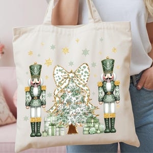May include: A cream-colored canvas tote bag featuring a festive graphic print of two green nutcrackers standing on either side of a decorated Christmas tree with a large bow on top, surrounded by gold glitter snowflakes and gift boxes.
