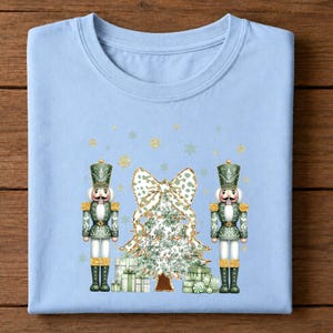 May include: A light blue t-shirt featuring a graphic print of two green nutcrackers standing on either side of a Christmas tree topped with a large leopard print bow. The design includes stacked gift boxes and scattered gold glitter snowflakes.