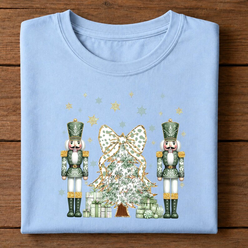 May include: A light blue t-shirt featuring a graphic print of two green nutcrackers standing on either side of a Christmas tree topped with a large leopard print bow. The design includes stacked gift boxes and scattered gold glitter snowflakes.