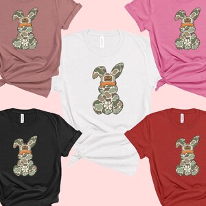 May include: Five t-shirts in different colours featuring a camouflage bunny rabbit wearing a hat. The bunny is in the centre of each shirt. The shirts are pink, white, black, red and brown.