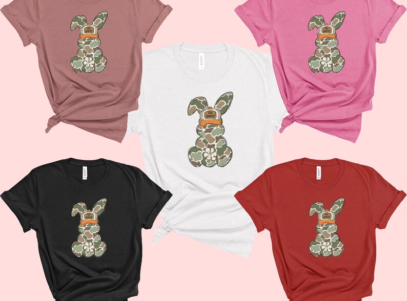 May include: Five t-shirts in different colours featuring a camouflage bunny rabbit wearing a hat. The bunny is in the centre of each shirt. The shirts are pink, white, black, red and brown.