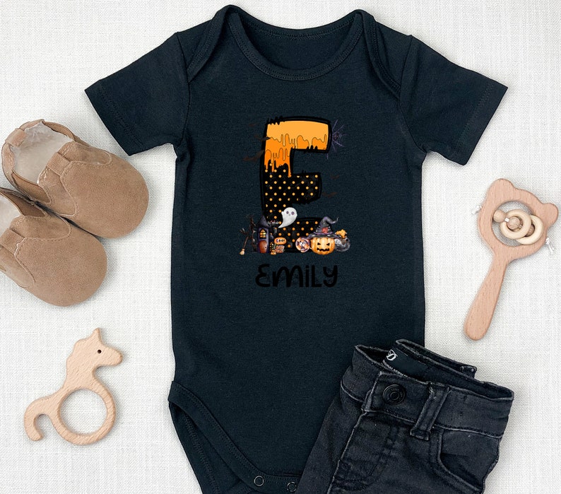 May include: Black baby bodysuit with a Halloween-themed design. The letter "E" is decorated with orange dripping paint and black polka dots. Below the letter is a collection of Halloween icons, and the name "EMILY" is printed below.