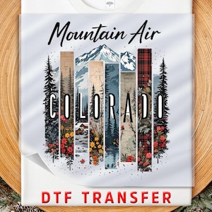 May include: White t-shirt with a colorful graphic featuring the text "Mountain Air Colorado." The design includes mountain scenery, trees, and floral elements within vertical stripes. The words "DTF TRANSFER" are at the bottom.