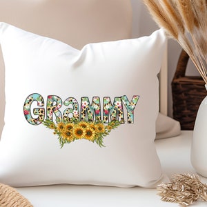 Op de afbeelding: Een wit kussen met het woord "GRAMMY" in zwarte omrande letters. De letters zijn gevuld met een bloemen- en luipaardprint. Een bloemenkrans van zonnebloemen bevindt zich onder het woord.