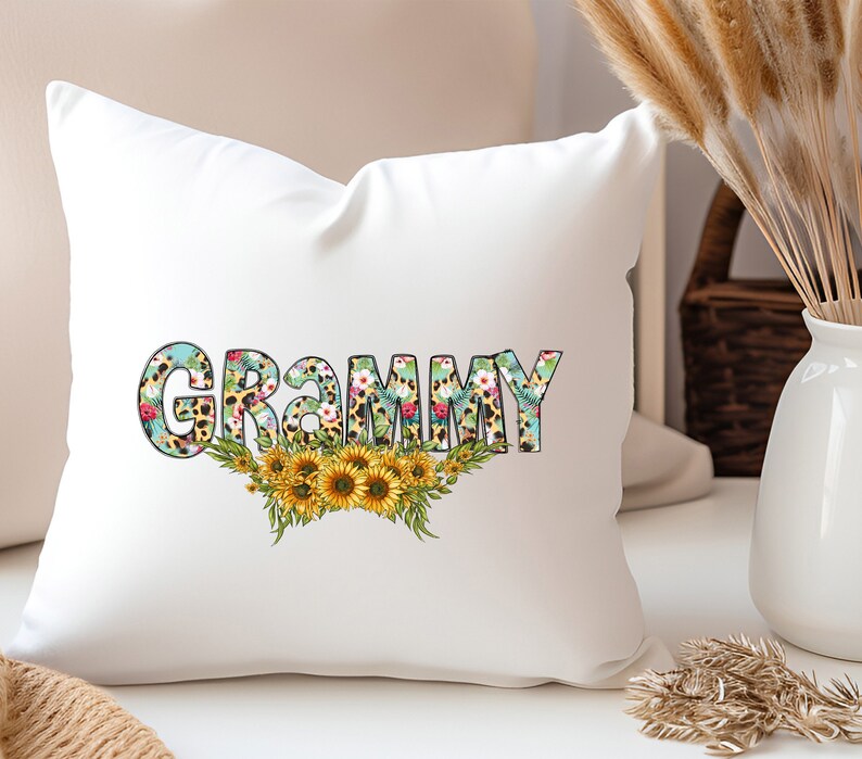 Op de afbeelding: Een wit kussen met het woord "GRAMMY" in zwarte omrande letters. De letters zijn gevuld met een bloemen- en luipaardprint. Een bloemenkrans van zonnebloemen bevindt zich onder het woord.