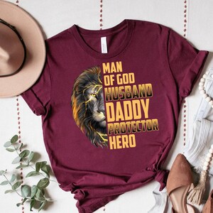 May include: A maroon t-shirt with a lion graphic and the text "Man of God, Husband, Daddy, Protector, Hero" in gold lettering.