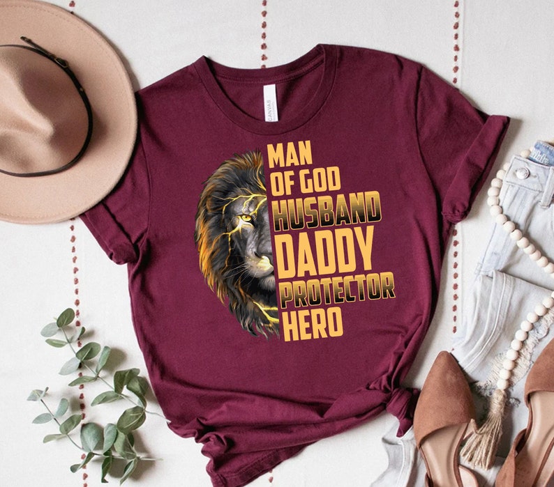 May include: A maroon t-shirt with a lion graphic and the text "Man of God, Husband, Daddy, Protector, Hero" in gold lettering.