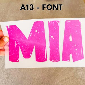 May include: A heat transfer decal on a white backing sheet featuring the name MIA in large, bold, hot pink capital letters. The letters have a distressed, textured finish with white highlights and the text A13 - FONT printed above the decal.