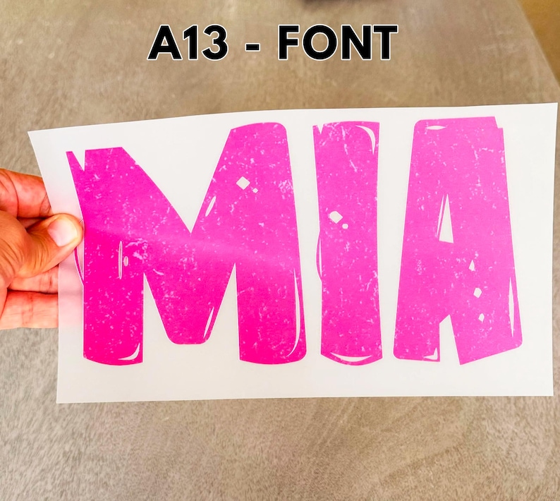 May include: A heat transfer decal on a white backing sheet featuring the name MIA in large, bold, hot pink capital letters. The letters have a distressed, textured finish with white highlights and the text A13 - FONT printed above the decal.
