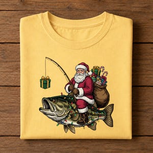 May include: A yellow t-shirt featuring a graphic of Santa Claus riding a large bass fish. Santa wears a red suit, carries a brown sack of gifts, and holds a fishing rod with a green gift box on the line. The fish is decorated with colorful lights.