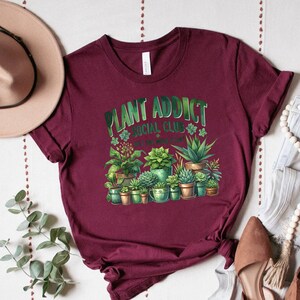 May include: Burgundy t-shirt with a green graphic of plants in pots and the text "Plant Addict Social Club - It's one more plant!"