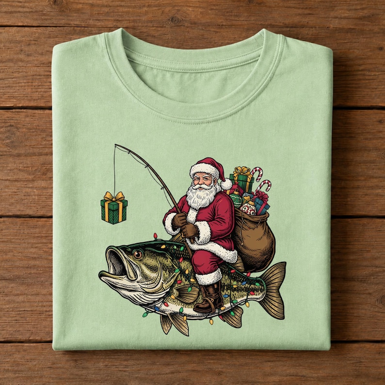 May include: A light green t-shirt featuring a graphic of Santa Claus riding a large fish. Santa wears a red suit, carries a brown sack filled with wrapped gifts, and holds a fishing rod with a green gift box dangling from the line.