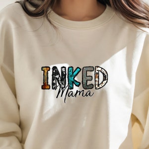 以下が含まれることがあります： カラフルな模様の文字で「INKED Mama」と書かれた白いスウェットシャツです。