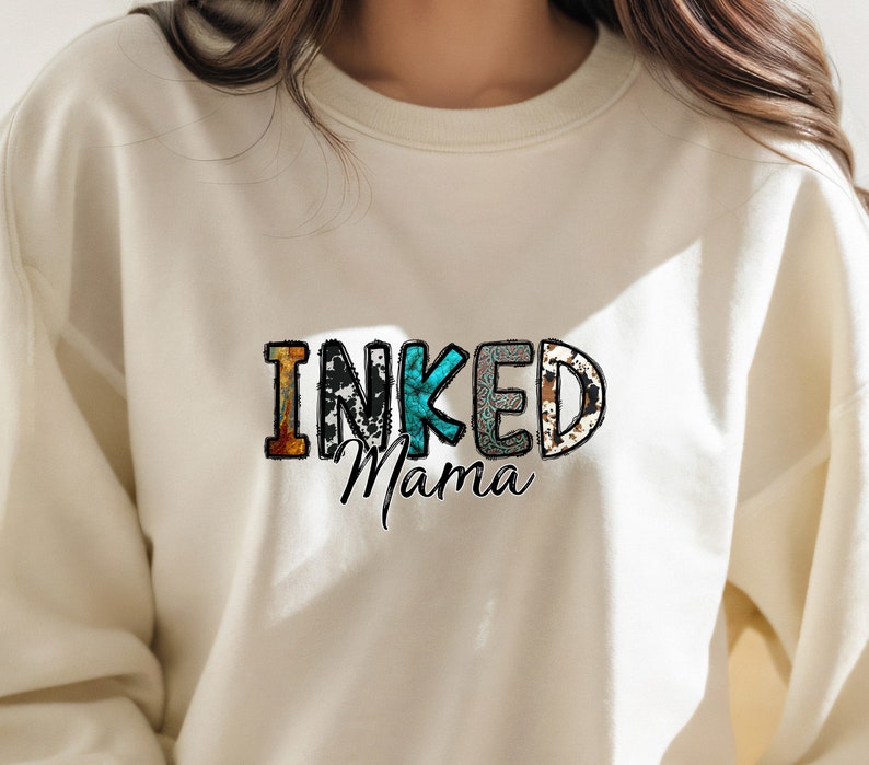 以下が含まれることがあります： カラフルな模様の文字で「INKED Mama」と書かれた白いスウェットシャツです。