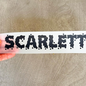 May include: A black vinyl decal on a white backing sheet featuring the name SCARLETT in a bold, dripping, liquid-like font style. The letters have rounded edges and small circular accents scattered around the characters to create a melting effect.