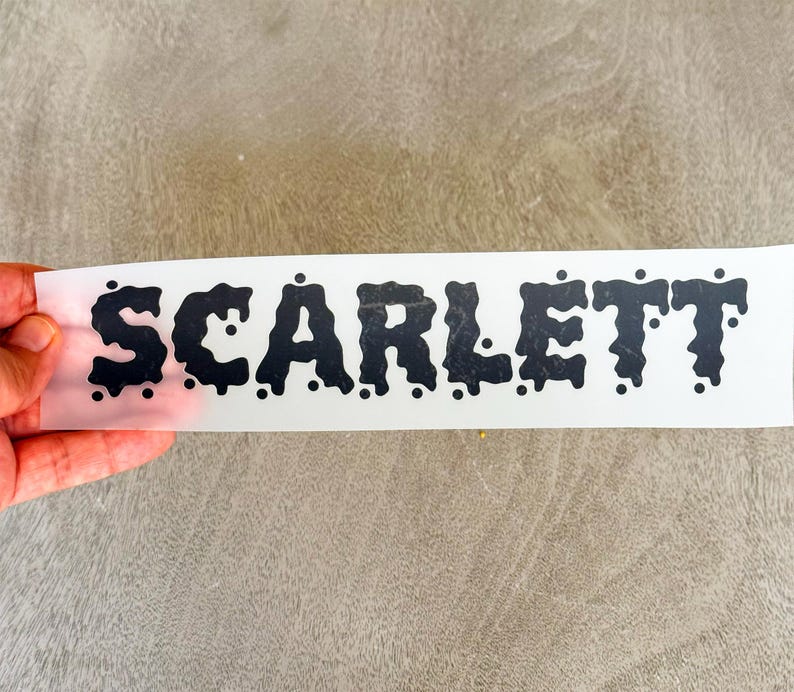 May include: A black vinyl decal on a white backing sheet featuring the name SCARLETT in a bold, dripping, liquid-like font style. The letters have rounded edges and small circular accents scattered around the characters to create a melting effect.