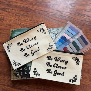 Könnte beinhalten: Zwei weiße Canvas-Reißverschlusstaschen mit schwarzem Text, der "Be Wary Be Clever Be Good" lautet.