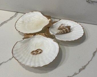 Puede incluir: Tres platos blancos en forma de concha con ribete dorado. El plato de la izquierda tiene un anillo de oro en su interior. El plato de la derecha tiene dos conchas en su interior.