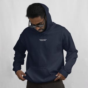 Puede incluir: Una sudadera con capucha azul marino con un bolsillo delantero y una capucha. La palabra "tipper" está bordada en blanco en el pecho. La sudadera está hecha de un material suave.