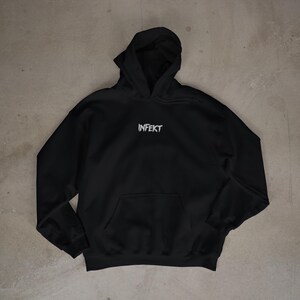 Puede incluir: Sudadera negra con bolsillo delantero y capucha. La palabra "INFEKT" está impresa en blanco en el pecho. La sudadera está hecha de un material suave y es adecuada para ropa casual.
