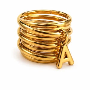 May include: A gold-colored ring with a stackable design. The ring features a dangling letter "A" charm. The ring is made of a shiny, metallic material and is set against a white background.