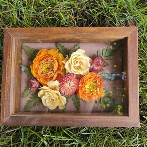 May include: A rectangular wooden frame encases a floral arrangement. The arrangement features vibrant orange and yellow flowers, with hints of pink and green foliage. The frame is set against a backdrop of green grass.