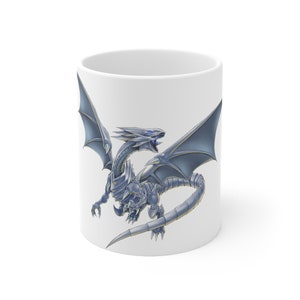 Puede incluir: Taza de cerámica blanca con una ilustración detallada de un dragón azul y plateado. El dragón tiene alas extendidas, garras y dientes afilados, ojos azules y una larga cola. La taza es de tamaño estándar.