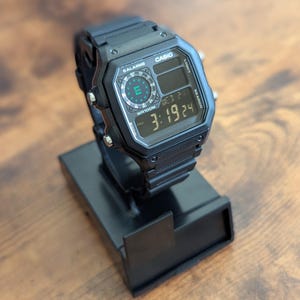 May include: Black Casio digital watch with a rectangular face, displaying the time 3:19:24. The watch has buttons labeled "Light" and "Search". The watch is on a black stand.