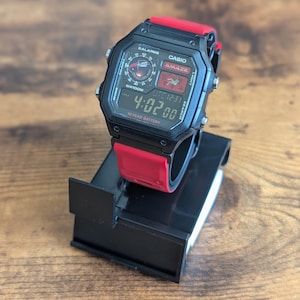 Peut inclure: Une montre num&eacute;rique Casio Amaze noire avec un bracelet rouge est pr&eacute;sent&eacute;e sur un support noir. L'&eacute;cran de la montre affiche l'heure 4:02:00, avec le texte "WR100M" et "10 YEAR BATTERY" visible. La montre a plusieurs boutons et un affichage num&eacute;rique.