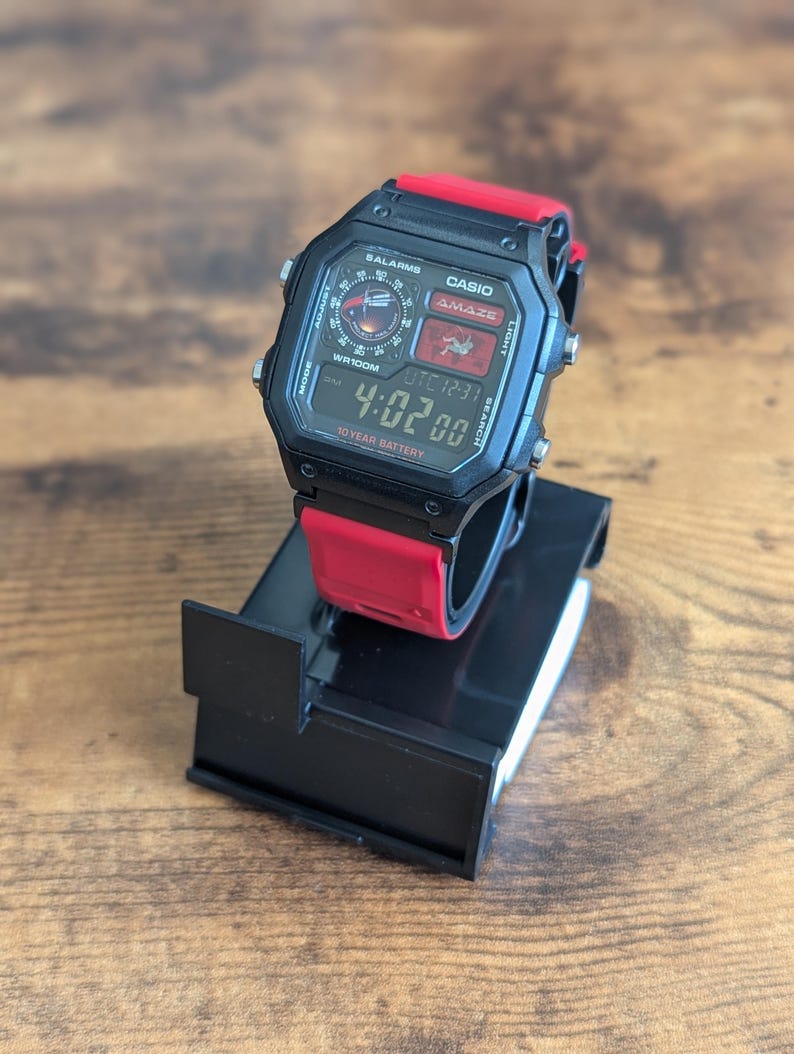Peut inclure: Une montre num&eacute;rique Casio Amaze noire avec un bracelet rouge est pr&eacute;sent&eacute;e sur un support noir. L'&eacute;cran de la montre affiche l'heure 4:02:00, avec le texte "WR100M" et "10 YEAR BATTERY" visible. La montre a plusieurs boutons et un affichage num&eacute;rique.