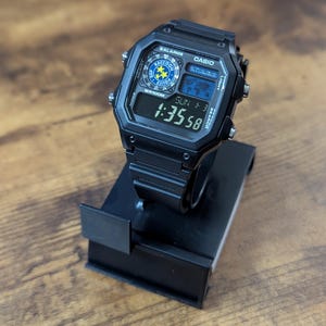 May include: Black Casio digital watch with a square face, displayed on a black stand. The watch face shows the time, date, and a world map graphic. The watch has the text "BALARMS", "CASIO", and "WR100M".